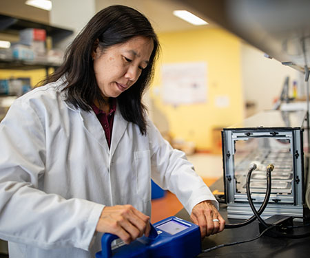 A researcher wearing a white lab coat operates air monitoring equipment in a laboratory, part of a project studying how to improve indoor air quality and reduce airborne health risks.